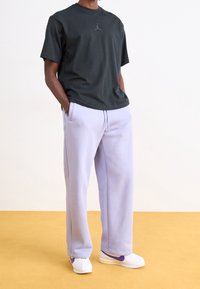 Osoba ubrana w czarną koszulkę Jordan, jasnofioletowe dresy i białe tenisówki z fioletowymi akcentami stoi na żółtej podłodze.