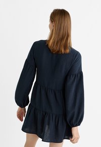 Vestido azul marino con un diseño en capas, mangas largas abullonadas y una tela texturizada. La parte trasera presenta un escote redondo y detalles de costura visibles.