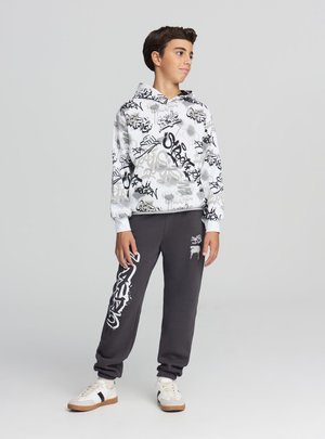 Ragazzo adolescente in piedi, indossa una felpa bianca con cappuccio con stampa graffiti, pantaloni della tuta scuri con scritte bianche su una gamba e scarpe da ginnastica bianche con strisce nere.