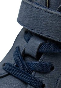 Scarpa in pelle blu scuro con superficie testurizzata, dotata di una fascia in Velcro e lacci intrecciati. Ingrandimento che mette in evidenza i dettagli delle cuciture.