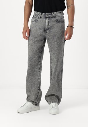 Grå, falmede denim cargo bukser med brede ben, fem lommer og synlige sting. Har en teksturert overflate og frontglidelås. Brukt med hvite sneakers.