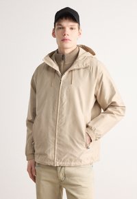 Beige huva jacka med dragkedja fram, elastiska ärmslut och sidfickor. Lättviktsmaterial, slät textur, har ett tonalt mönster. Bärs över en beige topp.