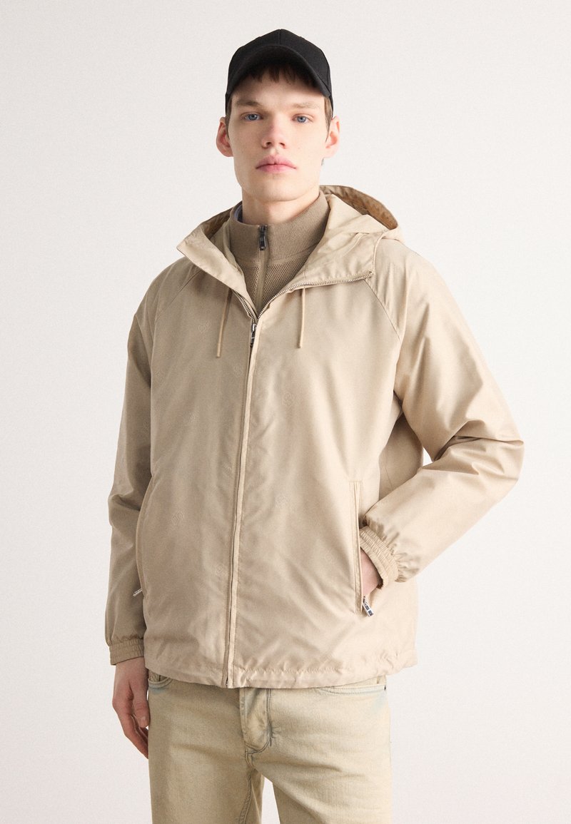 Beige huva jacka med dragkedja fram, elastiska ärmslut och sidfickor. Lättviktsmaterial, slät textur, har ett tonalt mönster. Bärs över en beige topp.