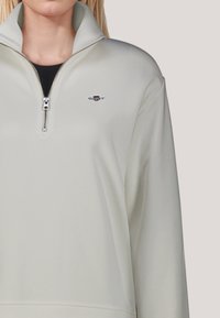 Felpa crema con zip e colletto alto, con un piccolo logo sul petto sinistro e texture liscia e morbida.