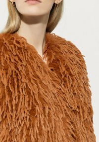 Künstlicher Pelzmantel in einem satten Braunton mit einer zerzausten Textur und einem tiefen Ausschnitt. Verfügt über ein weiches, mehrschichtiges Design für zusätzliche Wärme.