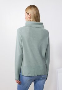 Green knit sweater with a high collar, long sleeves, and a textured finish. The hem is slightly longer at the back, paired with blue jeans.