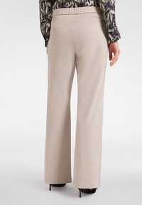 Beige wijde beenbroek met een zachte textuur en een elastische tailleband, gecombineerd met zwarte hoge hakken. Achteraanzicht toont schone lijnen.