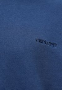Blauer Strickstoff mit einer subtilen gerippten Textur und einem gestickten Carhartt-Logo in Dunkelblau, das leicht versetzt ist.