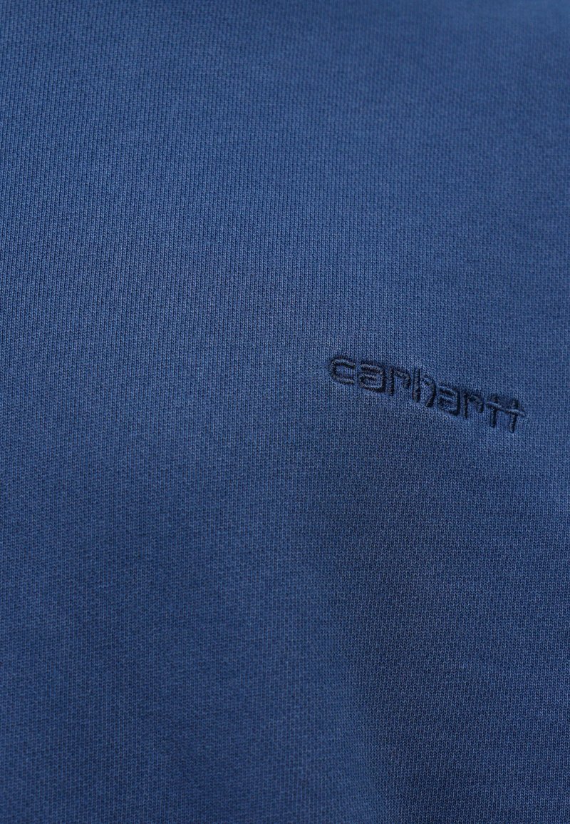 Blauer Strickstoff mit einer subtilen gerippten Textur und einem gestickten Carhartt-Logo in Dunkelblau, das leicht versetzt ist.
