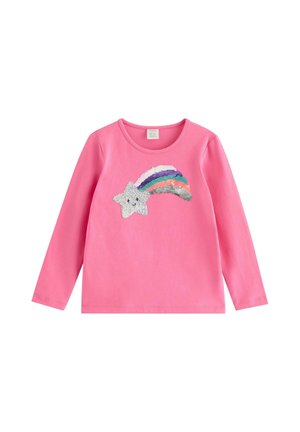 Barn iført lyserød kasket, lyserød langærmet trøje med regnbuedesign, blå jeans med stjernepatches, stående med hænderne på hoften.