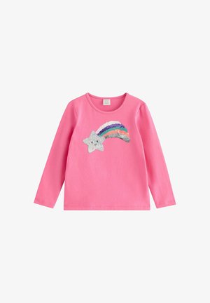 Barn iført lyserød kasket, lyserød langærmet trøje med regnbuedesign, blå jeans med stjernepatches, stående med hænderne på hoften.