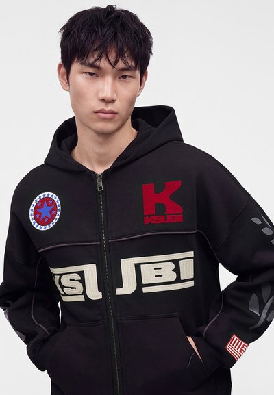 Jeune homme aux cheveux noirs portant un sweat à capuche noir avec des patches et logos rouges, blancs et bleus, mains dans les poches, expression neutre.