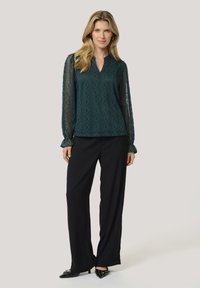 Blusa in pizzo turchese con maniche lunghe trasparenti, scollatura a V, motivi geometrici; abbinata a pantaloni neri a gamba larga e scarpe nere a punta.