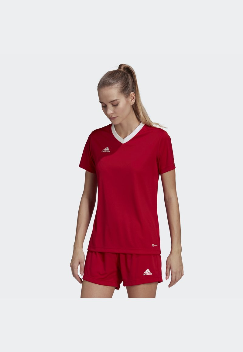 Punainen urheiluasu, jossa on pyöreä kaula-aukko ja valkoiset yksityiskohdat t-paidassa sekä yhteensopivat shortsit. Valmistettu kevyestä ja hengittävästä kankaasta. Adidas-logo näkyvillä.