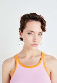 Roze geribbelde tanktop met oranje afwerking, met een ronde hals. Model draagt zilveren hoopoorbellen. Effen witte achtergrond.