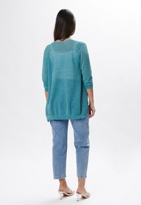 Cárdigan de punto turquesa con mangas tres cuartos, diseño texturizado y dobladillo trasero redondeado. Combinado con jeans azul claros y tacones blancos.