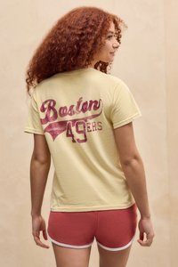 Žena s kučeravými červenými vlasmi, oblečená v žltom tričku "Boston 49ers" a červených športových kraťasoch, stojí na pozadí v beige farbe.