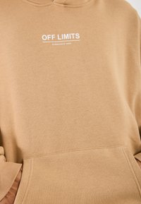 Sudadera beige con bolsillo frontal y texto en blanco "OFF LIMITS" subrayado, y texto más pequeño "DANGEROUS AREA" debajo.