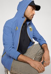 Μπλε ελαφρύ μπουφάν με κουκούλα, χρυσές λεπτομέρειες και κεντημένο λογότυπο. Διαθέτει τσέπες με φερμουάρ και αδιάβροχο ύφασμα.