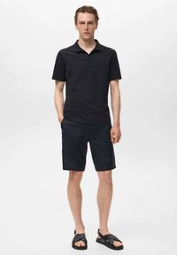 Svart polo skjorta med korta ärmar, matchad med mörka shorts och svarta sandaler. Tyget verkar lätt och mjukt.