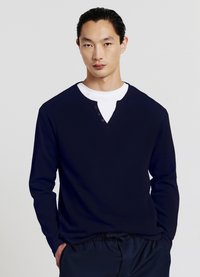 Chemise bleu marine à manches longues et col en V avec un tissu texturé, comportant trois boutons marron au niveau du col et un t-shirt blanc visible en dessous.