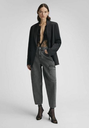 Mujer con blazer negro, blusa de encaje negro transparente, jeans cargo negros sueltos de tiro alto, calcetines negros transparentes y zapatos negros de tacón alto.