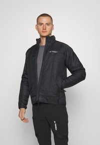 Chaqueta negra aislante con un diseño acolchado, cuello alto, cierre frontal con cremallera, dos bolsillos laterales y el logo "TERREX" en el pecho.