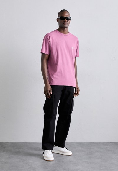 Camiseta de algodón rosa con cuello redondo y detalle de logo, combinada con pantalones cargo negros. Zapatillas blancas completan el atuendo. Lleva puestas gafas de sol.