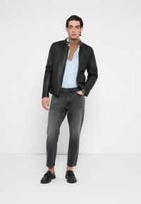 Zwarte leren bomberjack, lichtblauw overhemd, bruine coltrui, grijze spijkerbroek en zwarte instapschoenen. Model staat met handen in zakken.