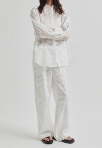 Osoba w białej, perforowanej koszuli z długim rękawem i pasujących szerokich spodniach, w czarnych sandałach, stojąca na jednolitym tle.