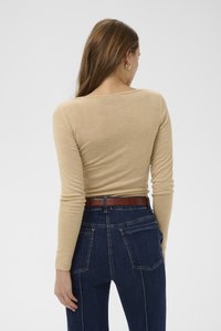 Hosszú ujjú, bézs színű felső kerek nyakkivágással, sötétkék farmerrel kombinálva, amely barna texturált övvel és jól látható hátsó zsebekkel rendelkezik.