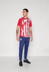 Mann steht und trägt ein rot-weiß gestreiftes Atlético Madrid Fußballtrikot und blaue Sporthosen mit Teamlogos, schwarze Turnschuhe und sichtbare Tattoos.