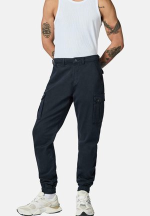 Mann trägt schwarze Cargo-Joggerhose, weißes Tanktop, weiße Sneakers und sichtbare Arm-Tattoos und steht mit den Händen hinter dem Rücken.