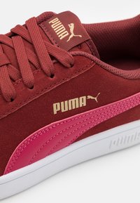 Punainen mokkanahkainen lenkkari, jossa vaaleanpunaiset nahkaiset yksityiskohdat, kultainen "PUMA"-logo, nauhallinen malli ja valkoinen kumipohja. Pinta on teksturoitu ja siinä on ompeludetaljeja.