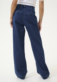 Vaalean siniset leveälahkeiset farkut korkealla vyötäröllä, joissa on kaksi takataskua ja ommellut helmat, näyttävät takaa.