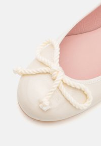 Sapatilha em material sintético brilhante de creme, com bico arredondado e um laço de corda branca como detalhe. O forro interior é de um tom rosa suave.