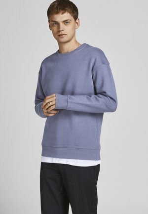 Sudadera lila claro con cuello redondo, puños y dobladillo de canalé, hecha de material suave. Superpuesta a una camiseta blanca. Pantalones negros.