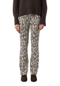 Dolna część ciała w spodniach z nadrukiem wężowym, ciemnobrązowym swetrze i ciemnobrązowych lordach stojąca na białym tle.