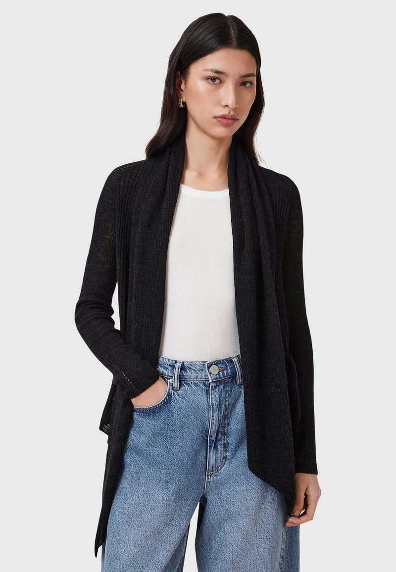 Black ribbed cardigan with open front, long sleeves, and draped collar, worn over a white top and high-waisted blue denim jeans.