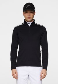 Svart långärmad zip-up topp med vita accentremsor på axlarna, tillverkad av ett slätt material. Kombinerad med vita byxor och en svart keps.