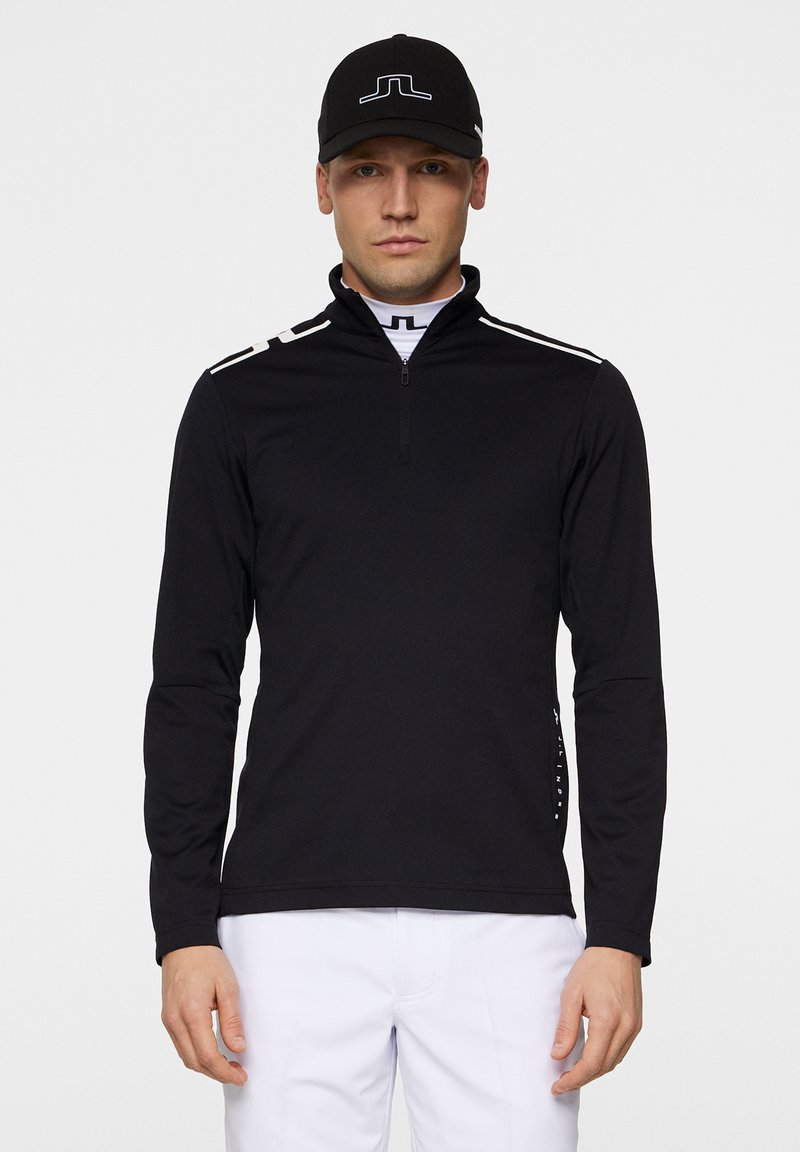 Svart långärmad zip-up topp med vita accentremsor på axlarna, tillverkad av ett slätt material. Kombinerad med vita byxor och en svart keps.