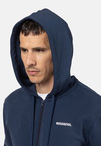 Marinefarbene Zip-Hoodie aus weichem Material mit Kapuze mit Kordelzug und weißem Logo-Text auf der Brust. Verfügt über eine glatte Textur und eine taillierte Passform.