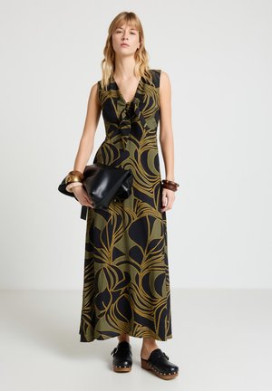 Donna che indossa un maxi vestito senza maniche nero e verde oliva con motivo a fantasia, tiene una grande pochette nera, con braccialetti di legno e zoccoli neri.