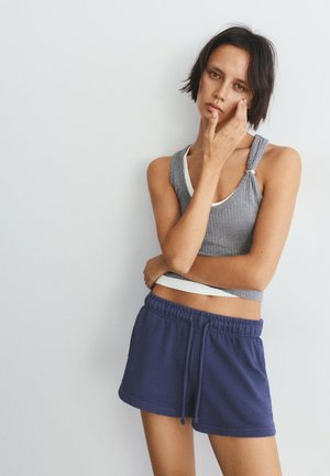 Mladi žena s kratkom tamnom kosom, odjevena u sivu majicu bez rukava i tamnoplave kratke hlače, dira lice, stoji uz jednobojni svijetli zid.