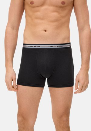 Torso masculino vistiendo bóxers ajustados negros con cintura gris que lleva el texto "YSABEL MORA", de pie contra un fondo claro y sencillo.