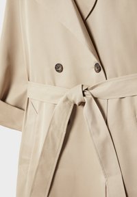 Beige trenchcoat met dubbele knopen, brede revers, zijzakken en een taille met riem die vooraan in een knoop is gestrikt.