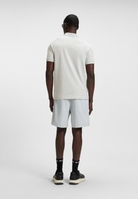 Man wearing a light beige polo shirt, light gray shorts, black socks with white text, and black sneakers, standing facing away on a white background.