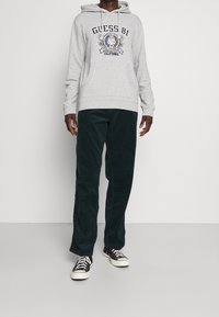Sudadera de algodón gris con el logo "GUESS 81", combinada con pantalones de pana verde oscuro y zapatillas negras con suelas blancas.