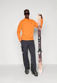 Oranžinė ilgarankovė palaidinė, juodos slidinėjimo kelnės su kišenėmis, juodos slidinėjimo batai ir slidės su reguliuojamomis tvirtinimo detalėmis, pateikti prieš neutralią fono spalvą.