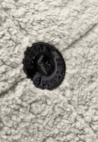 Zwarte ronde knop met de tekst "Frieda & Freddies" op een textuur van gebroken wit wollen stof, close-up.